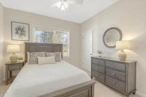 A bedroom with a wooden bed, white bedding, two nightstands with lamps, a dresser with a round mirror above it, and a window with blinds letting in natural light. Light-colored walls and carpet flooring.