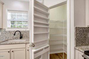 A kitchen with white cabinets, a granite countertop, a black faucet, and a window above the sink. An open door reveals a pantry with white wire shelves and wood flooring.