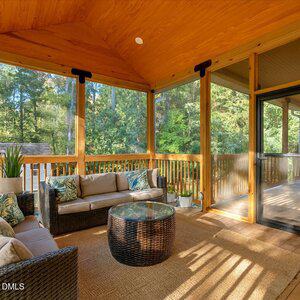 A screened-in porch features wicker furniture with beige cushions, a round glass coffee table, potted plants, and a wooden ceiling. Sunlight enters through the screens, revealing a view of trees outside.