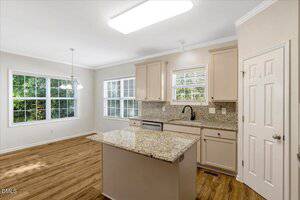 Bright kitchen with light beige cabinets, granite countertops, a central island, stainless steel dishwasher, wood flooring, and large windows offering views of greenery outside. A light fixture hangs over the dining area.