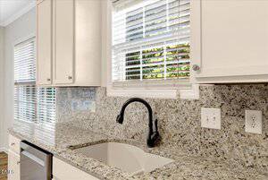A kitchen sink with a black faucet is set in a speckled granite countertop. White cabinets, backsplash, and multiple electrical outlets are visible. There are two windows with white blinds above and next to the sink.