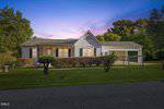 Charming single-story house at 208 W Fleming Street, Creedmoor, with light gray siding, front porch, attached carport, and a well-maintained lawn bordered by trimmed bushes and a chain-link fence, set against a sunset sky with trees behind.