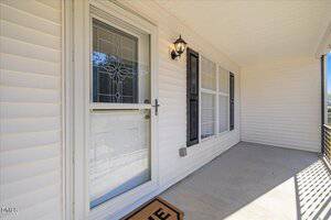 A covered front porch with cream-colored siding, concrete flooring, a glass storm door with decorative design, a lantern-style wall light, black shutters, and large windows. A doormat is partially visible.