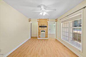 A bright room with light wood flooring, cream-colored walls, a white ceiling fan, and a large window. A section of wall features wood accents and a small white vent or heater. Two doorways lead to other rooms.