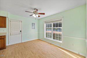 A room with light green walls, a large window with white blinds, a door with a window, light wood flooring, and a ceiling fan with lights. Sunlight is coming through the window onto the floor.