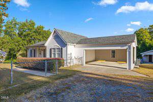 Single-story house with light gray siding, covered front porch, attached two-car carport, chain-link fence, and a driveway, set on a grassy lot with trees in the background under a blue sky.