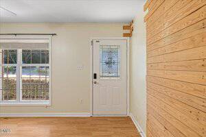 A room with light wood flooring, a white front door with decorative glass, a wall with horizontal wooden planks, and a large window with blinds on the left.