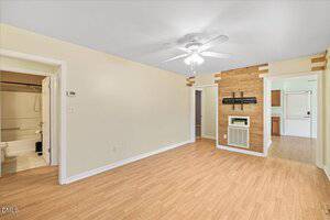 A living room with light wood flooring, beige walls, a ceiling fan with lights, and a wooden accent wall around a vent. Three doorways lead to a bathroom, a hallway, and a kitchen with visible cabinetry.