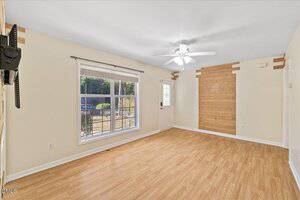 A bright, empty living room with light wood flooring, cream-colored walls, a ceiling fan with light, a large window, a wall-mounted TV bracket, a door, and a wood accent wall panel.