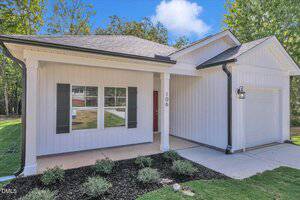 A single-story house with white vertical siding, black shutters, a covered front porch, and a one-car garage. The house has a front lawn with small shrubs, and the number 106 is visible near the entrance.