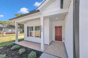 A white house with vertical siding has a covered front porch, a red front door, and the number 106 on a support column. There are small bushes along a mulched area by the entrance.