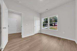 A small, empty room with light-colored wood flooring, white walls, two closed doors, and two large windows showing a view of a yard and neighboring houses.