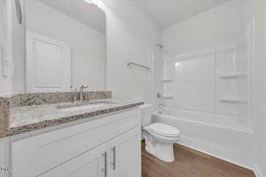 A bathroom with a white vanity and granite countertop, a large mirror, a toilet, and a bathtub with an integrated shower and shelving. The floor is a light wood material and the walls are painted white.