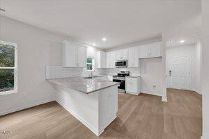 A modern kitchen with white cabinets, stainless steel appliances, light granite countertops, subway tile backsplash, and light wood flooring. The space includes a breakfast bar and is adjacent to an entryway with a white door.