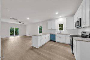 Modern open-concept kitchen and living area with light wood floors, white cabinets, stainless steel appliances, and large windows. Sliding glass door in the living area opens to a green outdoor space.