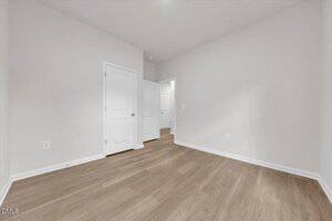 A small, empty room with white walls, light wood flooring, a closed white door, an open white door, two visible electrical outlets, and a white baseboard trim. The room is well-lit with no furniture.