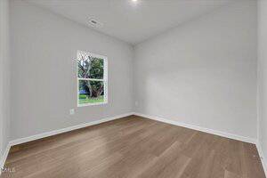 A small, empty room with light gray walls, a single window showing a tree and grass outside, and light brown wood flooring. There are no furnishings or decorations in the room.