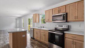 A modern kitchen with light wood cabinets, stainless steel appliances, white countertops, a center island, and large windows letting in natural light. The space has wood-look flooring and white walls.