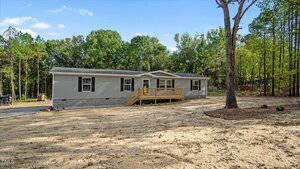 This single-story manufactured home at 212 Ellington Path, Henderson features beige siding and a wooden front porch on a cleared lot surrounded by trees. The yard offers mostly bare soil and minimal landscaping.