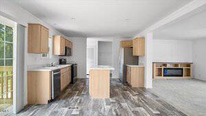 A modern kitchen with light wood cabinets, stainless steel appliances, an island in the center, and a wood-look floor. An open living area with built-in shelving and carpet is visible on the right. Sliding glass doors are on the left.