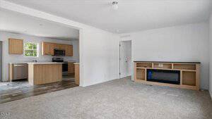 A modern open-concept living room with light carpet, a built-in entertainment unit, and an adjacent kitchen featuring wood cabinets, stainless steel appliances, and a center island. The walls are painted white.