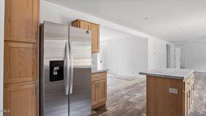 A modern kitchen with stainless steel refrigerator, wooden cabinets, an island with a light countertop, and a view into an empty, open living area with wood-look flooring.