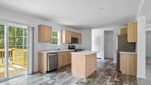 Modern kitchen with light wood cabinets, stainless steel appliances, a small island, and sliding glass doors leading to a deck. The space has white walls, large windows, and wood-style flooring.