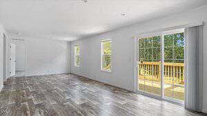 A spacious, empty room with wood-patterned flooring, white walls, two large windows, and a sliding glass door leading to a wooden deck outside. Natural light fills the room.
