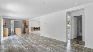 Open-plan living space with wood-look flooring, white walls, and recessed lighting. Visible kitchen area with light wood cabinets and island. Doorway leads to bathroom and carpeted room. Large windows allow natural light.