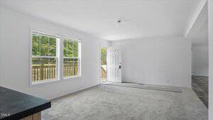 A bright, unfurnished living room with light gray carpet, white walls, two large windows, a door to the outside, and a doormat near the entryway. Sunlight streams in from the windows.