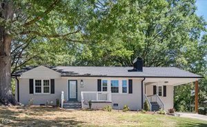 A single-story house at 304 Lanier Street, Oxford, features a light gray brick exterior, dark roof, small covered porch with white railing, attached carport, and a minimally landscaped yard shaded by large trees.