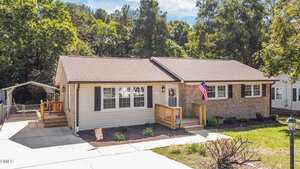 Charming single-story house at 111 Ward Avenue, Oxford, with light-colored siding and brick accents, a front porch with steps, an American flag, a paved driveway, surrounding trees, and a small shed or carport to the left.