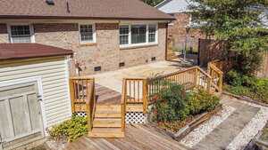 A backyard with a wooden deck and stairs leading to a concrete patio. The patio is adjacent to a brick house with three windows. There are garden beds with shrubs and a shed on the left side.