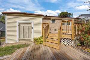 A wooden deck with stairs leads up to a raised entryway at the back of a brick house. There is a small shed or storage area with a weathered wooden door to the left of the deck. The sky is partly cloudy.