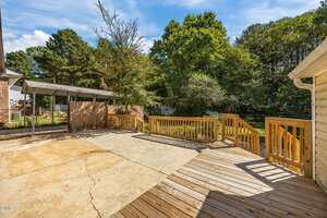 A backyard area with a concrete patio, a wooden deck with railings, a carport, and surrounding trees in the background on a sunny day.