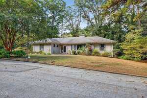 Single-story house with a gray roof and light-colored exterior, surrounded by trees and shrubs. The front yard has patchy grass and a driveway with visible cracks in the foreground.
