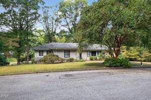 Single-story light gray house with a dark roof, surrounded by mature trees and shrubs, sits on a slightly sloped lawn next to a paved street under a clear sky.