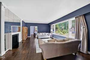 A spacious living room with dark blue walls, hardwood floors, two beige sofas, a white area rug, large windows with patterned curtains, a wooden bookshelf, and a fireplace with a mirrored wall above it.