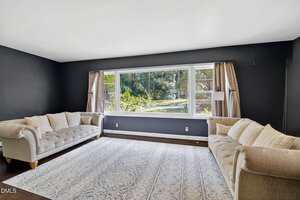 A living room with dark walls, two tufted beige sofas facing each other, a large patterned area rug, and a wide window with beige curtains overlooking greenery outside. Two wall-mounted lamps frame the window.