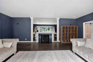 A spacious living room with dark blue walls, a white fireplace centered between two columns, a large mirror above it, two light-colored couches, a wooden shelving unit, and a patterned area rug on a dark wood floor.