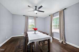 A dining room with light gray walls, dark wood floors, a ceiling fan, two windows with patterned curtains, and a rectangular table covered with a white tablecloth and a vase of red flowers, surrounded by six chairs.