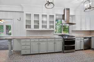 A spacious kitchen features light gray cabinets, a patterned tile floor, a gas stove with a vent hood, glass-front upper cabinets, a tile backsplash, and a window overlooking a green yard.