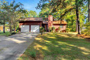 This charming single-story house at 378 Daniel Boone Trail in Henderson features a brown exterior, two-car garage, chimney, and sloped roof, surrounded by trees and a sun-dappled lawn. A concrete driveway leads to the garage.