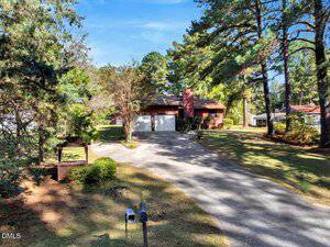 A single-story brick house with a two-car garage sits at the end of a curved driveway, surrounded by tall trees and a spacious, grassy yard in a suburban neighborhood.