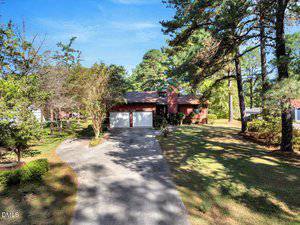 A single-story house with a two-car garage is surrounded by trees and shrubs. A curved driveway leads from the street to the garage. The lawn and landscaping are well maintained under a clear sky.