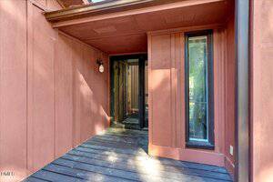 A covered entryway with reddish-brown wooden siding, a dark wood floor, a single wall-mounted light fixture, and a glass door with sidelights leading to the interior. Sunlight casts shadows on the surfaces.