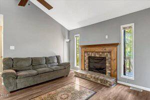 A gray living room with a dark green upholstered sofa, a wooden fireplace with tile detailing, two tall narrow windows, a floor lamp, and wood flooring with a small area rug.