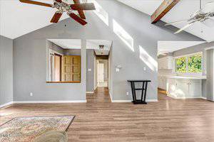 A spacious, bright room with wood flooring, gray walls, exposed wooden ceiling beams, ceiling fans, large windows, and an open view into a white kitchen and hallway with a front door.