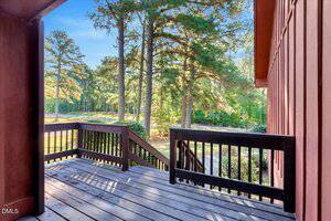A wooden deck with railing overlooks a yard with green grass and tall pine trees. Stairs lead down from the deck to the ground, and sunlight filters through the trees in the background.
