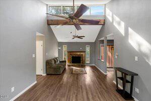 A living room with gray walls, wood flooring, a green sofa, a fireplace with a rug in front, ceiling fans, exposed wood beam, and upper windows letting in natural light. A small black table is against the right wall.